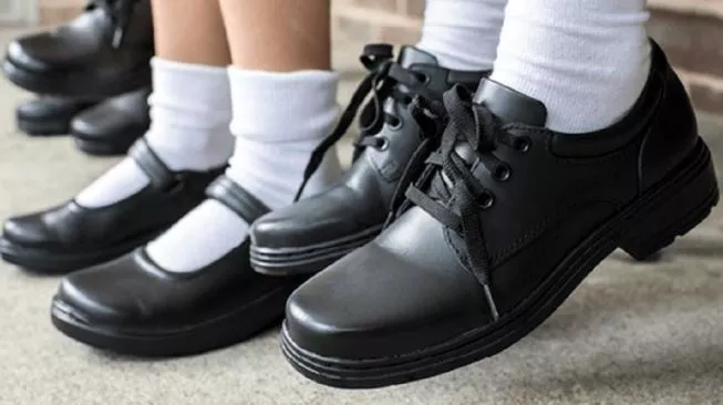 Close-up of a student wearing black leather lace-up shoes with white socks, with another pair in the background on a concrete floor.