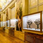 Museum gallery with glass display cases along a gold-toned wall, housing antique scientific instruments such as armillary spheres; ornate paintings and a frescoed ceiling above, and a tiled floor.