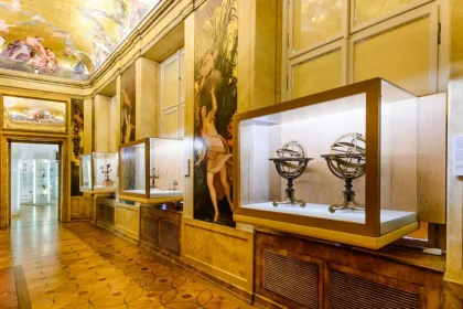 Museum gallery with glass display cases along a gold-toned wall, housing antique scientific instruments such as armillary spheres; ornate paintings and a frescoed ceiling above, and a tiled floor.