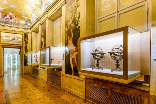 Museum gallery with glass display cases along a gold-toned wall, housing antique scientific instruments such as armillary spheres; ornate paintings and a frescoed ceiling above, and a tiled floor.