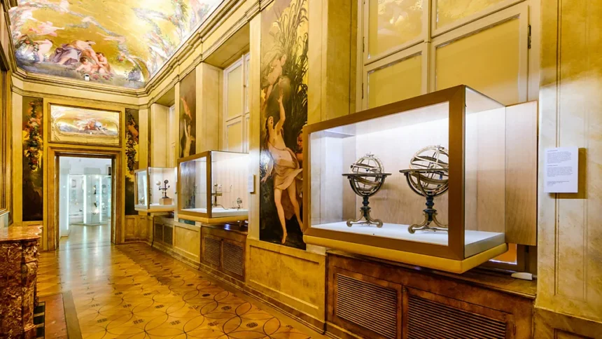 Museum gallery with glass display cases along a gold-toned wall, housing antique scientific instruments such as armillary spheres; ornate paintings and a frescoed ceiling above, and a tiled floor.