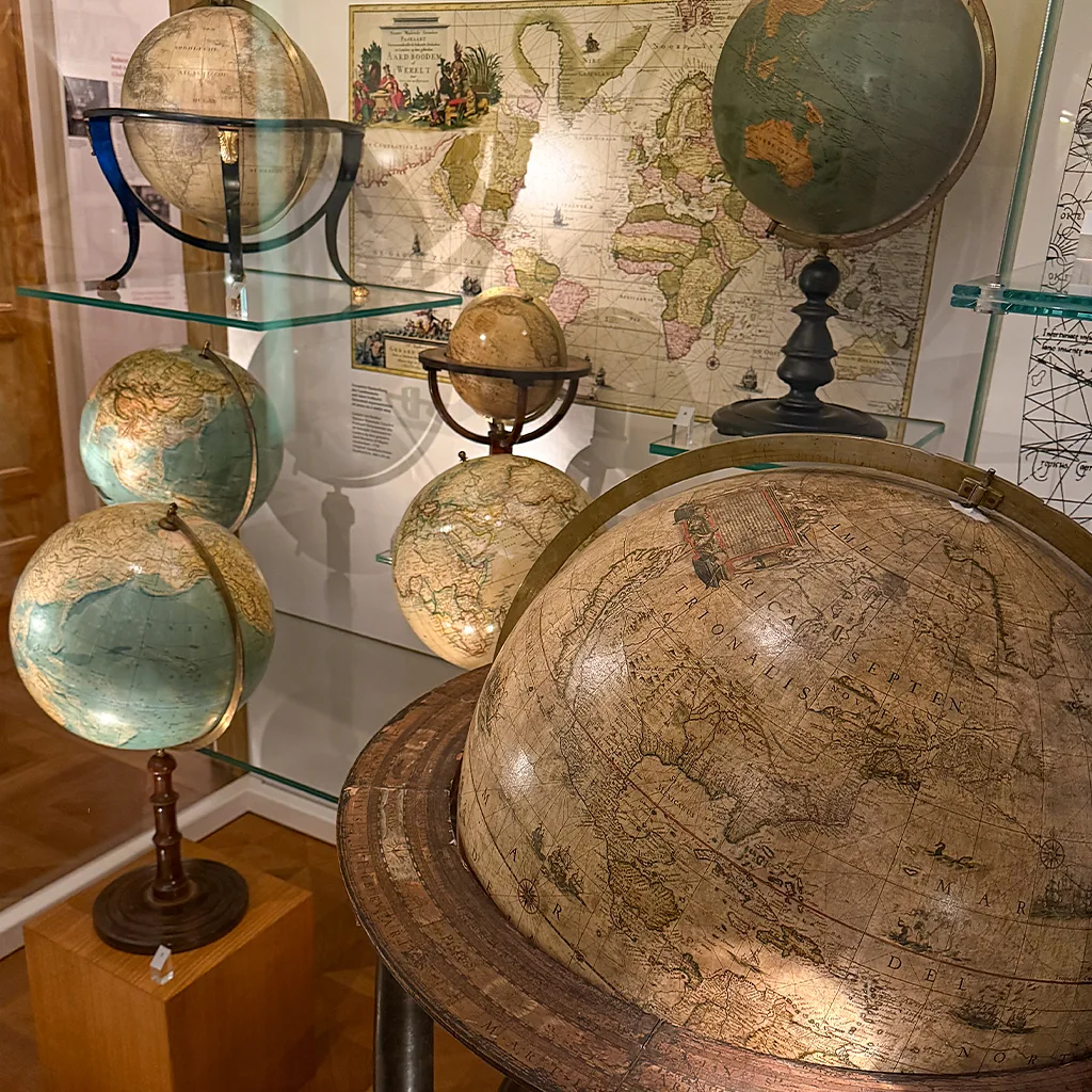 Group of antique globes on wooden stands in a museum display with a world map backdrop in the background.