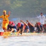 Dragon boat race: a yellow-clad paddler stands at the prow with arms raised, leading a team of paddlers through splashing water.