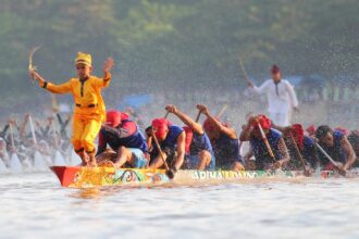 Dragon boat race: a yellow-clad paddler stands at the prow with arms raised, leading a team of paddlers through splashing water.