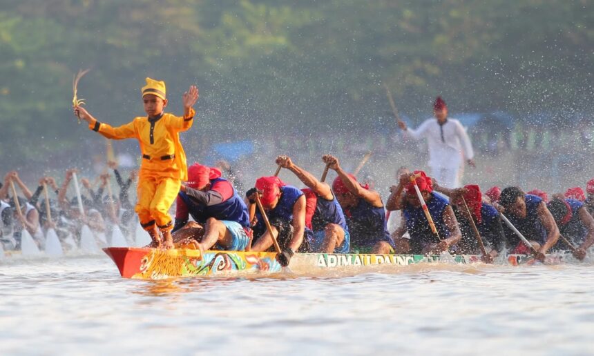 Dragon boat race: a yellow-clad paddler stands at the prow with arms raised, leading a team of paddlers through splashing water.