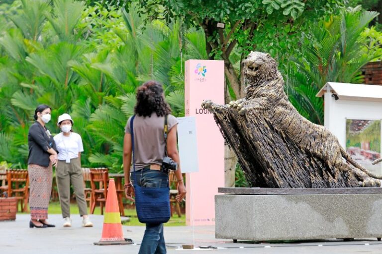Outdoor plaza with a large bear statue on a concrete pedestal and water flowing from its paws, set among lush tropical plants; several people wearing masks stand nearby.