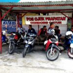 Six motorcycles parked under a red-roofed shelter with a group of men sitting on a bench behind them, near a signboard.