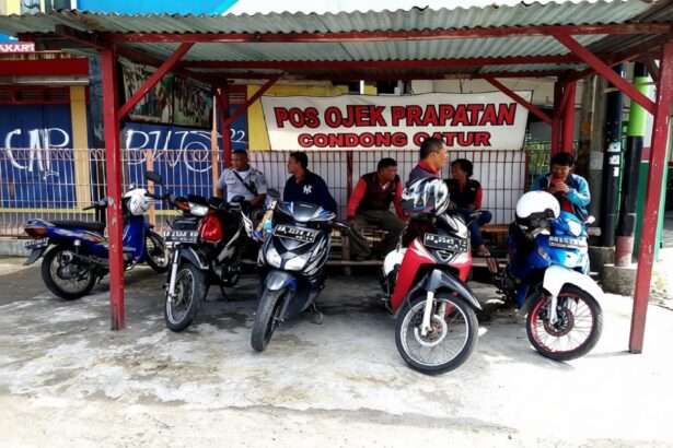 Six motorcycles parked under a red-roofed shelter with a group of men sitting on a bench behind them, near a signboard.