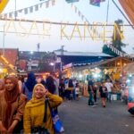 People posing under a colorful festival entrance that reads PASAR KANGEN at a busy night market.