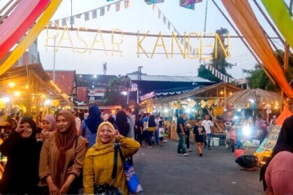People posing under a colorful festival entrance that reads PASAR KANGEN at a busy night market.