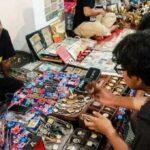 Vendor sits on the floor beside a colorful display of watches, smiling at a customer inspecting timepieces on a market stall table.