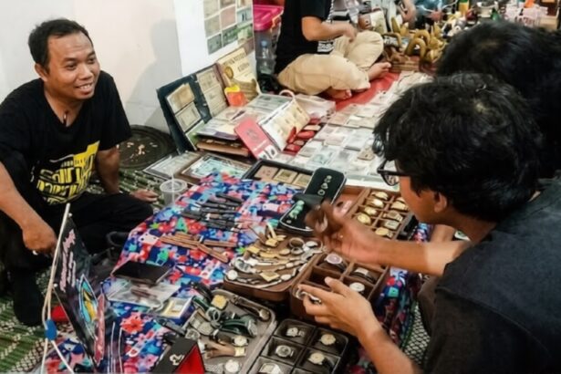 Vendor sits on the floor beside a colorful display of watches, smiling at a customer inspecting timepieces on a market stall table.