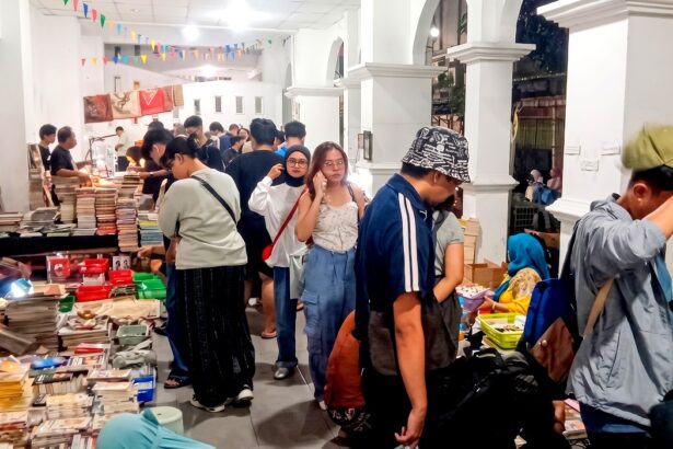 Crowded indoor book market with tables full of stacked books and shoppers browsing under colorful bunting ground overhead.