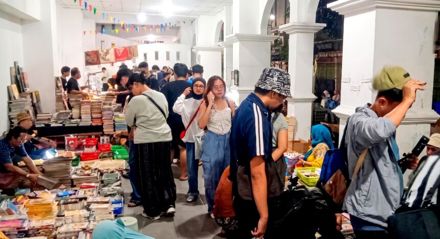 Crowded indoor book market with tables full of stacked books and shoppers browsing under colorful bunting ground overhead.