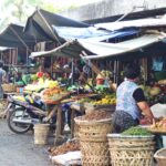 Outdoor street market scene with vendors under tarps selling fruits and vegetables; a man on a scooter passes by while produce baskets and piles of vegetables surround the stalls.
