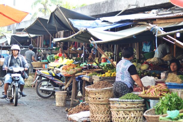 Outdoor street market scene with vendors under tarps selling fruits and vegetables; a man on a scooter passes by while produce baskets and piles of vegetables surround the stalls.