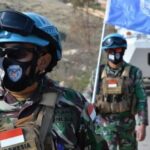 Indonesian peacekeepers in camouflage uniforms with blue helmets and masks walking along a road beside a UN vehicle with a blue UN flag in the background, showing Indonesian flags on their vests.