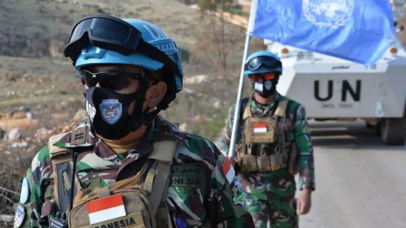 Indonesian peacekeepers in camouflage uniforms with blue helmets and masks walking along a road beside a UN vehicle with a blue UN flag in the background, showing Indonesian flags on their vests.