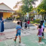 A woman in a blue dress and hijab stands in front of a group of children outdoors, leading an activity in a sunny courtyard with a yellow building in the background.