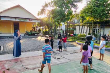 A woman in a blue dress and hijab stands in front of a group of children outdoors, leading an activity in a sunny courtyard with a yellow building in the background.