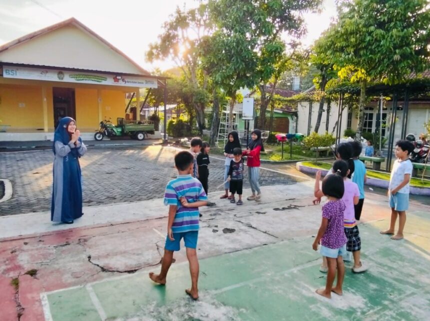 patok-lele-1024x765 - Mabur.co A woman in a blue dress and hijab stands in front of a group of children outdoors, leading an activity in a sunny courtyard with a yellow building in the background.