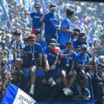 A blue-clad sports team sits on a decorated bus during a victory parade, waving to fans and wearing gold medals.