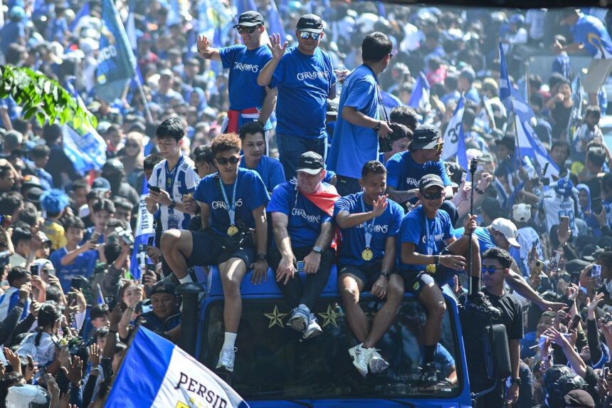 A blue-clad sports team sits on a decorated bus during a victory parade, waving to fans and wearing gold medals.