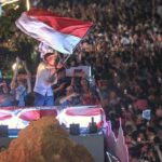 Man on a festive float waves a large red-and-white flag above a cheering crowd at night, fans taking photos.