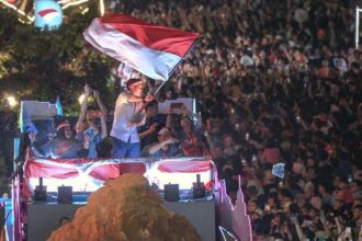 Man on a festive float waves a large red-and-white flag above a cheering crowd at night, fans taking photos.