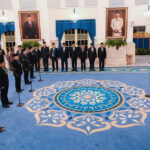 Formal ceremony in a grand hall with men in suits standing in rows and a speaker at a microphone, surrounded by portraits and blue carpet patterns.