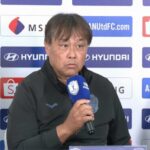 Middle-aged man in a dark jacket speaks into a blue microphone at a press conference, sponsor logos on the backdrop behind him.