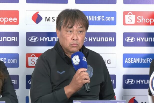 Middle-aged man in a dark jacket speaks into a blue microphone at a press conference, sponsor logos on the backdrop behind him.