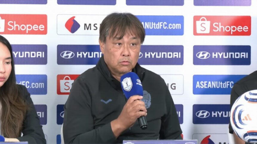 Middle-aged man in a dark jacket speaks into a blue microphone at a press conference, sponsor logos on the backdrop behind him.
