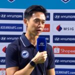 A man interviewed on a sports backdrop holding a blue Hyundai microphone, wearing a navy polo shirt with a badge.
