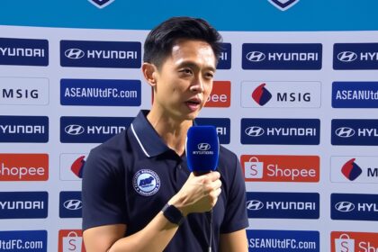 A man interviewed on a sports backdrop holding a blue Hyundai microphone, wearing a navy polo shirt with a badge.