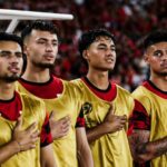 Four male soccer players in gold jerseys stand with hands over hearts during the national anthem in a stadium crowd behind them.