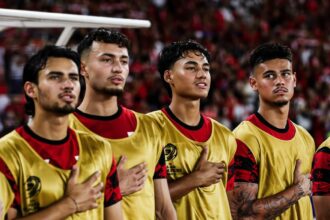 Four male soccer players in gold jerseys stand with hands over hearts during the national anthem in a stadium crowd behind them.