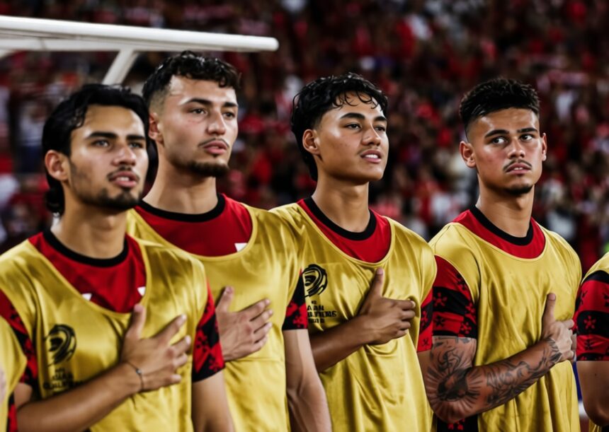 Four male soccer players in gold jerseys stand with hands over hearts during the national anthem in a stadium crowd behind them.