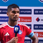 Soccer player in a red jersey gives a post-match interview, holding a blue microphone with a press backdrop behind him.