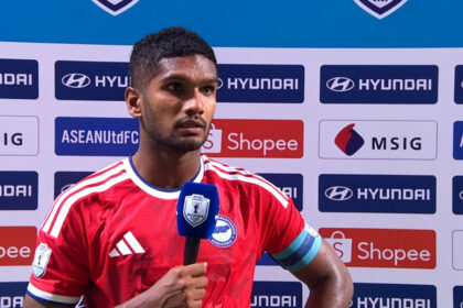 Soccer player in a red jersey gives a post-match interview, holding a blue microphone with a press backdrop behind him.
