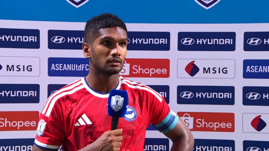 Soccer player in a red jersey gives a post-match interview, holding a blue microphone with a press backdrop behind him.