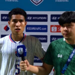Two young football players stand in front of sponsor logos; one holds a blue microphone, the other wears a green jersey with a lanyard.