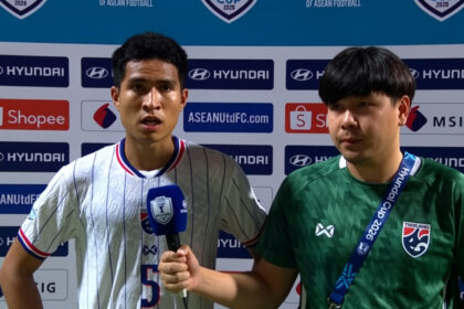 Two young football players stand in front of sponsor logos; one holds a blue microphone, the other wears a green jersey with a lanyard.