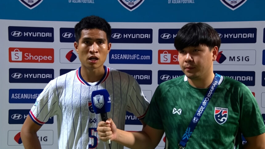 Two young football players stand in front of sponsor logos; one holds a blue microphone, the other wears a green jersey with a lanyard.
