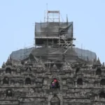 Stone temple wall with multiple niches and a central arched opening, covered in scaffolding for renovation on the upper levels.