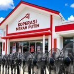 Front facade of a white building with red trim and a sign reading 'KOPERASI MERAH PUTIH' against a blue sky, with barbed wire in the foreground.