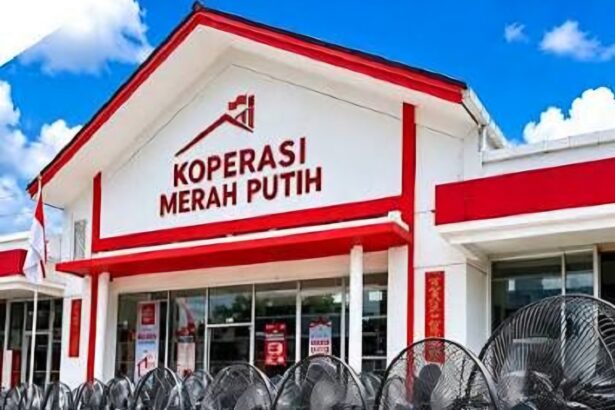 Front facade of a white building with red trim and a sign reading 'KOPERASI MERAH PUTIH' against a blue sky, with barbed wire in the foreground.