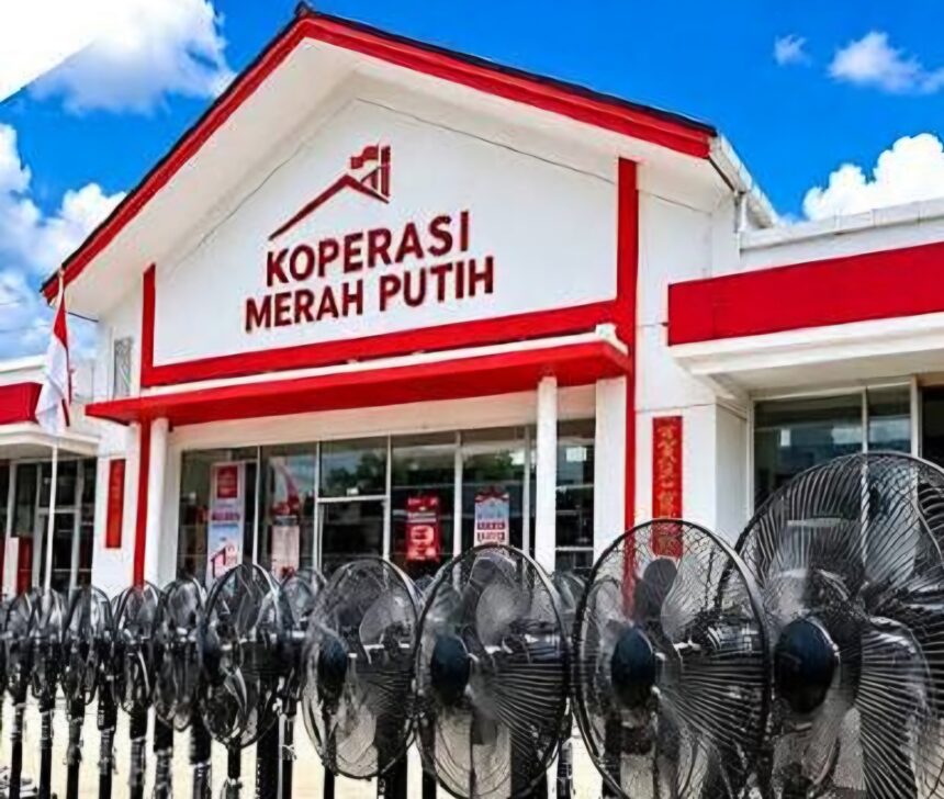 Front facade of a white building with red trim and a sign reading 'KOPERASI MERAH PUTIH' against a blue sky, with barbed wire in the foreground.