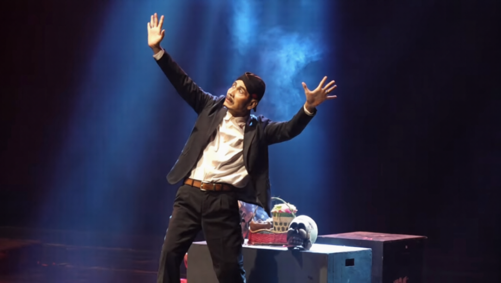 penggali kubur mulyono 2-ezremove - Mabur.co Man on a dark stage with both arms raised under blue spotlight, performing a theatrical gesture. A table with props sits behind him.