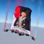 Skydivers hold up a large banner with a portrait of a male official in ceremonial dress, red-and-black flag colors, and Indonesian presidential text.